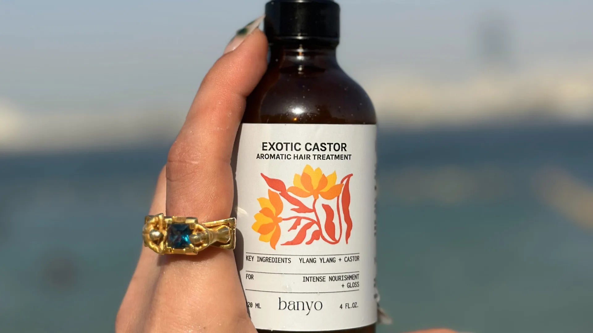 Hand holding a bottle of 'Exotic Castor Aromatic Hair Treatment' by banyo with a blurred beach background