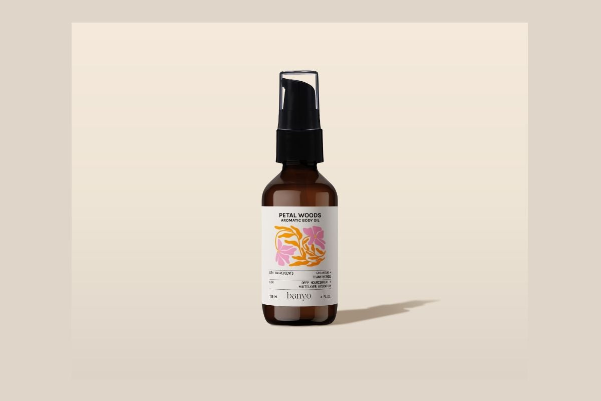 The Banyo Petal Woods Aromatic Body Oil, 4 oz amber pump bottle on a beige background.
