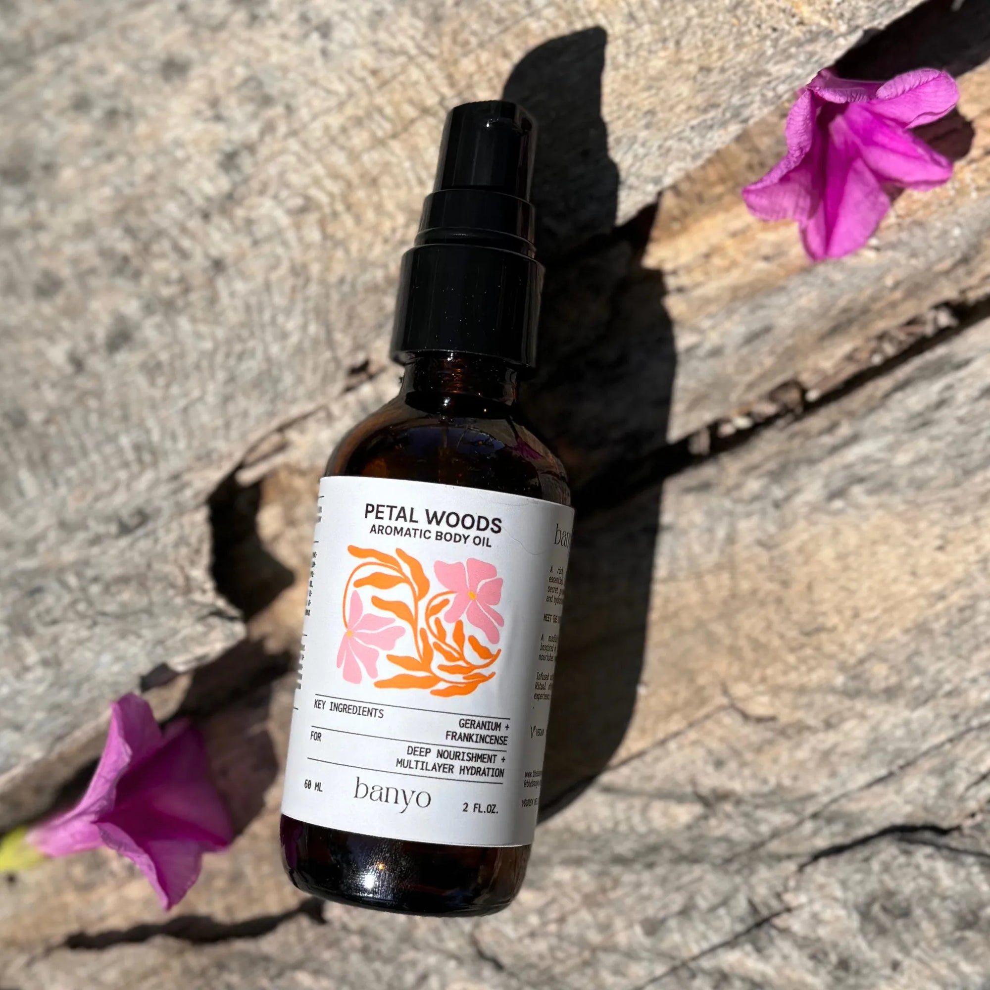 Bottle of Petal Woods Aromatic Body Oil on a stone surface with pink flowers.