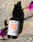 Bottle of Petal Woods Aromatic Body Oil on a stone surface with pink flowers.